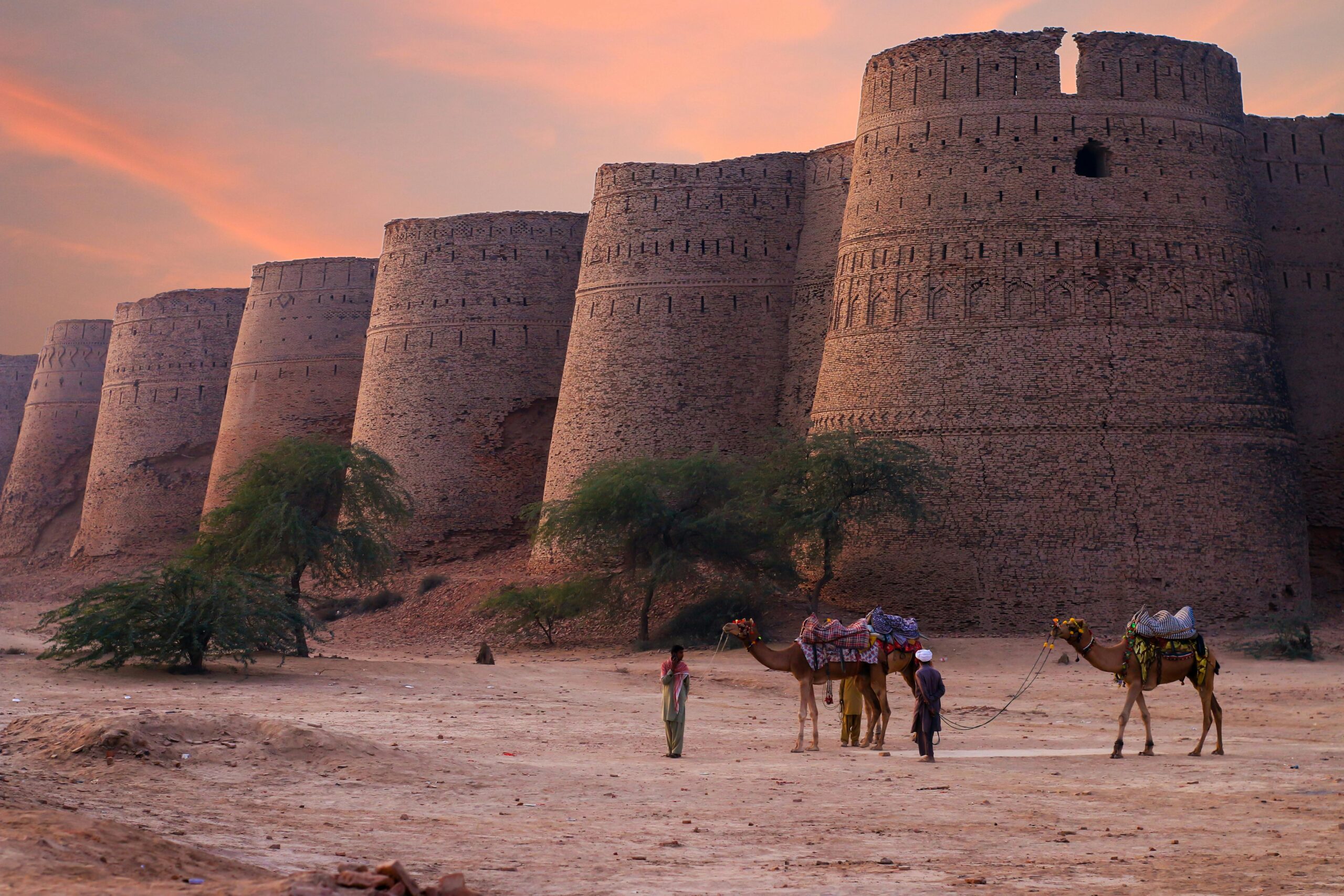 Scenic view of Derawar Fort's towers at sunset in the Punjab desert with camels and people.